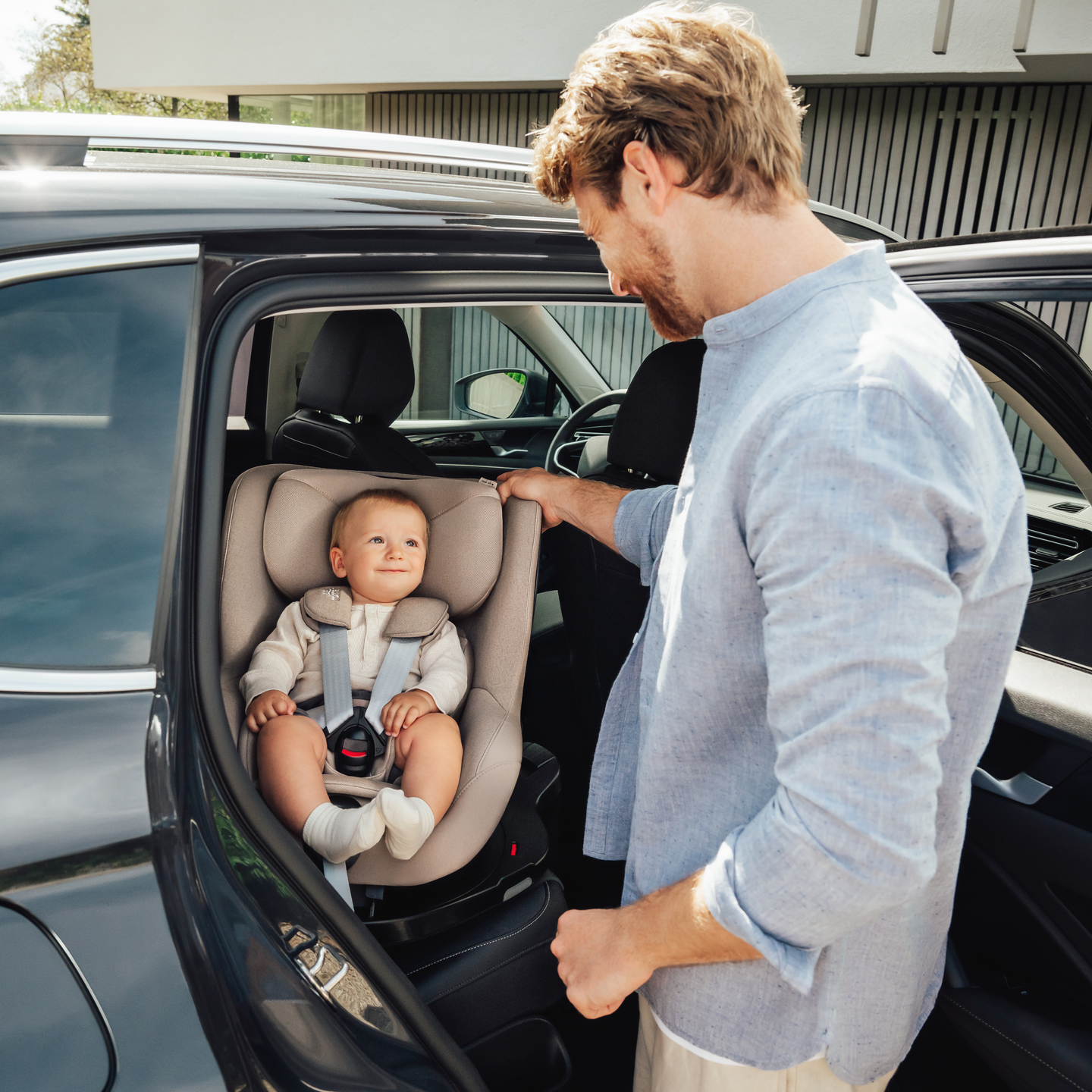 A smiling baby sitting safely in a DUALFIX PRO M in TEAK car seat, rear-facing in the back seat of a car, while an adult places the child inside.